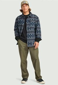 Man in een beige pet, blauw patroon overhemdjasje, zwart shirt, olijfgroene broek en zwarte sneakers die tegen een effen achtergrond staat.