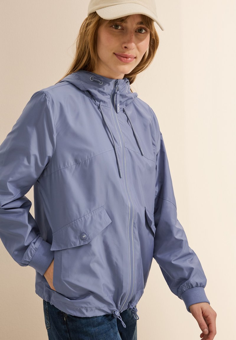 Veste zippée violette clair avec capuche, cordon de serrage, poignets élastiques et deux poches avant, présentant une texture lisse et brillante.