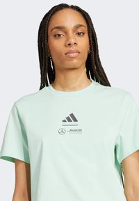 Mint zelena majica kratkih rukava izrađena od pamuka. Ima crni Adidas logo i Mercedes AMG Petronas brendiranje na prednjoj strani.
