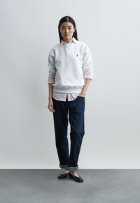 Νέα γυναίκα στέκεται με το ένα χέρι στην τσέπη, φοράει λευκό φούτερ πάνω από ροζ πουκάμισο, σκούρο παντελόνι με γυρισμένα μπατζάκια και μαύρες μπαλαρίνες, μπροστά σε απλό φόντο.