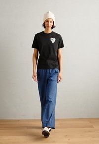 T-shirt nera a maniche corte con un design grafico a forma di cuore, abbinata a pantaloni denim larghi blu e a un berretto beige chiaro.