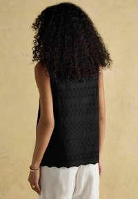 Melna bezvīļu blūze ar izšūtiem ziedu rakstiem un viļņotu apakšmalu. Izgatavota no viegla auduma, ar brīvu piegriezumu un gludu tekstūru.