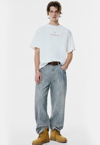Ung person står med den ene hånd i lommen, iført en oversized hvid T-shirt med rød tekst, løse lyseblå jeans, brunt bælte og tanstøvler.