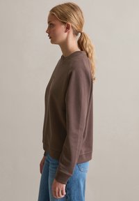 Sweatshirt marrone con scollatura a girocollo, polsini e orlo a costine, vestibilità casual, mostrato insieme a jeans in denim azzurro chiaro a vita alta.