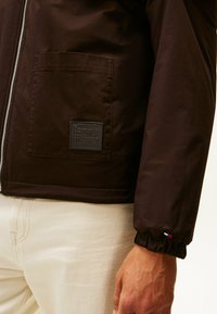 Brązowa kurtka z zamkiem błyskawicznym, z kieszonką naszywaną i gumową metką z logo. Rękawy z mankietami z trójkolorowym akcentem u dołu.