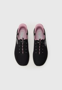 Juodos slip-on Skechers sportbačiai su rožine vidine apdaila ir dekoratyvinėmis raištelėmis, matomi iš viršaus ant baltos fono.