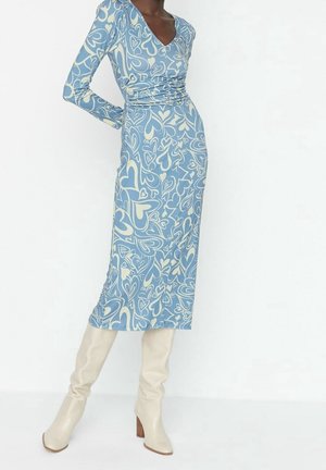 Kobieta ubrana w niebieską sukienkę midi z kremowymi wzorami serc, z długimi rękawami i dekoltem w serek, zestawioną z kremowymi kozaczkami sięgającymi kolan na słupku.