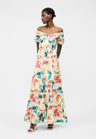 Γυναίκα με πολύχρωμο μακρύ φόρεμα με λουλούδια, έξω οι ώμοι και φουσκωτά μανίκια, στέκεται με τα χέρια πίσω από την πλάτη, κοιτάζοντας πλάγια.