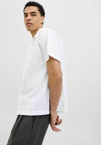 Camicia bianca a maniche corte con bottone frontale, caratterizzata da un motivo a griglia testurizzato. Indossata con pantaloni scuri, il modello posa di lato.