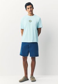 Hellblaues Baumwoll-T-Shirt mit einem bunten Grafikdruck auf der Brust, kombiniert mit Jeansshorts und hellgrauen Slippern.