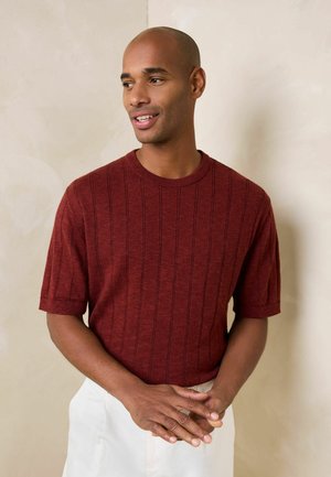 Man with short hair wearing a rust-colored, short-sleeve sweater and white pants, standing with hands clasped, looking to the side.