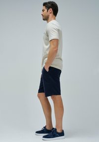 Camiseta de algodón beige, pantalones cortos azul marino, zapatillas de ante azul marino. Hombre de pie de lado, con las manos en los bolsillos, mostrando un atuendo casual.