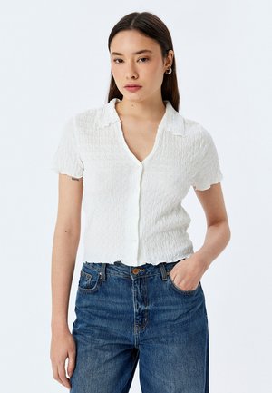 Witte T-shirt met korte mouwen, een gestructureerde, gekreukte afwerking, knoopsluiting aan de voorkant en een notch-kraag. Gecombineerd met hoge, blauwe jeans.
