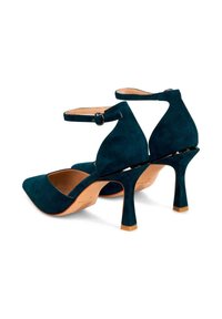 Scarpe con tacco alto in suede verde scuro, con punte appuntite, cinturini alla caviglia e tacchi curvi. La suola è di colore beige e presenta una texture liscia.