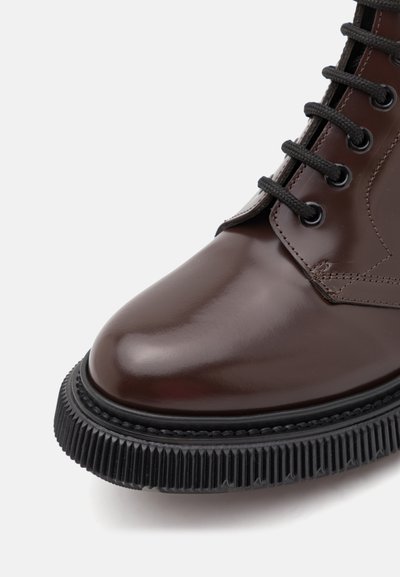 Bottines en cuir marron poli avec semelle en caoutchouc noir, bord texturé et six œillets noirs pour les lacets. Design de bout arrondi et lisse.