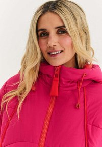 Jasně růžová zateplená bunda s vysokým límcem, kontrastním červeným zipem a stojánky, viditelným šitím a elastickými manžetami. Hladká textura.