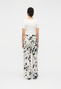 Λευκό τοπ με κοντά μανίκια, συνδυασμένο με παντελόνι φαρδιάς γραμμής που διαθέτει μαύρο και κρεμ μοτίβο πιτσιλιάς. Ομαλή υφή υφάσματος με ά νετη εφαρμογή.