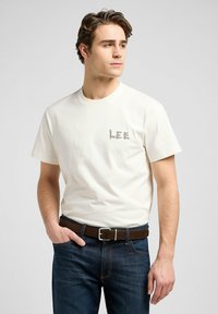 Vit bomullströja med grå "LEE"-logotyp på bröstet, ihop med mörk denimjeans och ett brunt bälte. Enkla rundhalsad design.