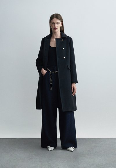 Frau steht und trägt einen langen schwarzen Mantel mit goldenen Knöpfen, schwarze Hosen mit weitem Bein und einem goldenen Kettengürtel, ein schwarzes Oberteil und weiße spitze Schuhe.