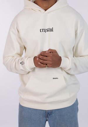 Mężczyzna ubrany w kremową bluzę z kapturem z napisem "crystal" na piersi i "dreams." na kieszeni, w połączeniu z niebieskimi dżinsami, ręce splecione na talii.