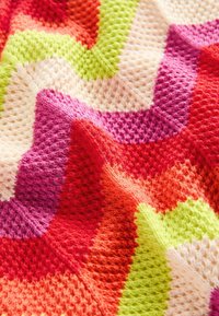 Multikleurige gebreide deken met een zigzagpatroon in oranje, roze, groen en crème. Zachte textuur met een gezellige uitstraling.