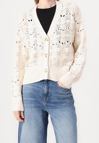 Stickad cardigan i krämfärg med en öppen, spetdesign. Har en V-ringning, tre stora knappar och ribbade ärmslut; kombinerad med blå denimjeans.