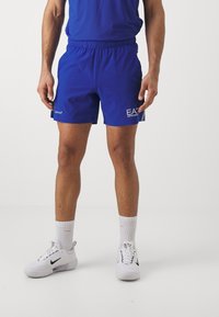Blå idrottsshorts med elastisk midja, prydda med "EA"-logotyp i vitt på sidan. Bärs tillsammans med vita Nike-sneakers och strumpor.