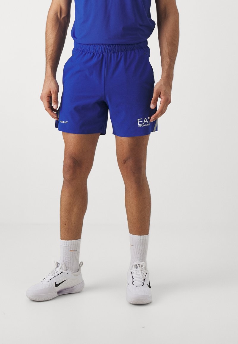 Blå idrottsshorts med elastisk midja, prydda med "EA"-logotyp i vitt på sidan. Bärs tillsammans med vita Nike-sneakers och strumpor.