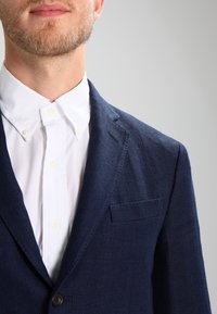 Man med ljus skägg som bär en vit knäppt skjorta och en marinblå kavaj mot en enkel grå bakgrund.