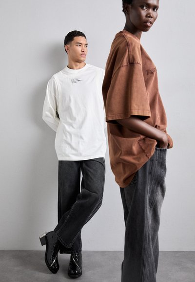 White long-sleeve shirt with a label, paired with brown short-sleeve shirt. Both wear dark jeans and glossy black heeled boots.