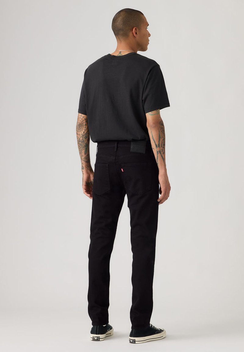 Homme tatoué portant un t-shirt noir, un jean noir et des baskets noires, debout, de dos, devant un fond gris uni.