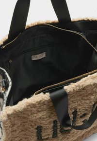Grande borsa a tote con un esterno beige texturizzato, dotata di un interno nero, chiusura con cerniera e manici in tessuto neri. Logo "LIU-JO" visibile.
