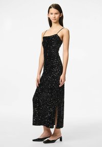 Crna večernja haljina s cvjetnim detaljima, tankim naramenicama, uskim siluetom, izrezom do sredine bedra i do koljena. Glatka tekstura; sjajna, reflektirajuća površina.