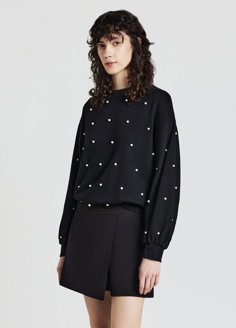 Maglione nero con perle rotonde in un motivo sparso, vestibilità ampia e maniche a palloncino, abbinato a una gonna nera aderente.