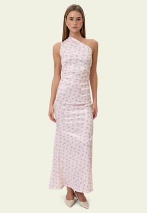 Moteris dėvi šviesiai rožinę, vieno peties maksi suknelę su mažais gėlių raštais ir dėvi smėlio spalvos aukštakulnius su smailiais galiukais, stovinti priešais paprastą foną.