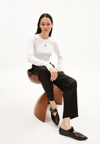 Weißes Langarmoberteil mit quadratischem Logo, kombiniert mit schwarzen maßgeschneiderten Hosen und schwarzen spitzen Ballerinas. Glattes Material und moderne Silhouette.