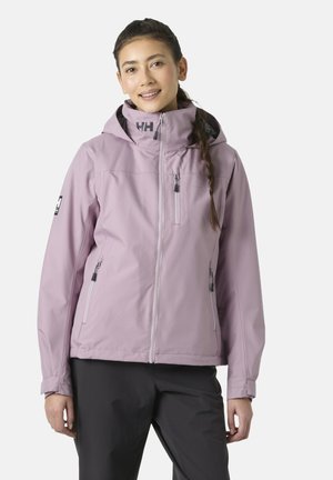Woman wearing a light purple hooded jacket with front and chest zip pockets and black pants, standing against a plain background.