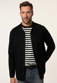 Cardigan nero con colletto e bottoni, sopra una camicia a righe bianche e nere. Design casual e aderente; dettaglio della tasca frontale e texture liscia.