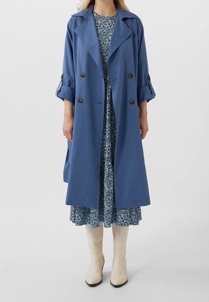 Kvinde iført blå dobbeltradet trenchcoat over en blå blomsterkjole med cremeknæhøje støvler, stående mod en ensfarvet grå baggrund.