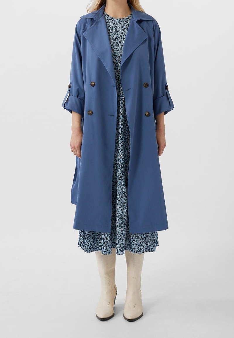 Femme portant un trench-coat bleu croisé par-dessus une robe fleurie bleue avec des bottes crème montantes jusqu'aux genoux, debout sur un fond gris uni.