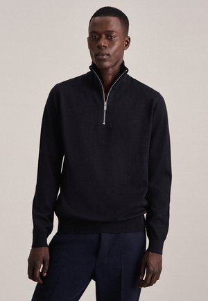 Maglione nero a maglia con zip a quarto di altezza, colletto alto, polsini e orlo a coste, e una consistenza liscia. Indossato sopra pantaloni scuri.