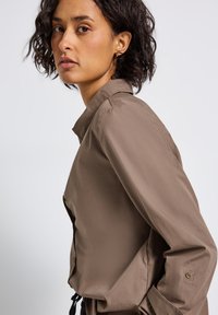 Donna con capelli ricci scuri che indossa una camicia marrone con bottoni, girata leggermente di lato contro uno sfondo chiaro e semplice.