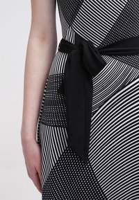 Svartvit mönstrad klänning med ränder och prickar. Inkluderar ett svart tygbälte knutet i midjan. Närbild av designen.