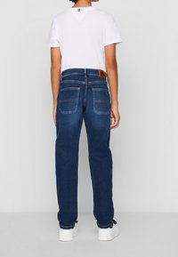 Jeans de mezclilla azul oscuro con corte recto, que presentan una parche de cuero marrón y dos bolsillos traseros. Combinados con una camiseta blanca y zapatillas blancas.