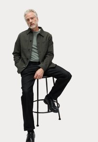 Dark green jacket with buttons, light green shirt, black pants, and black shoes. Sitting on a black stool against a white background.