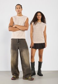 Haut sans manches beige tricoté avec un motif texturé ; mannequin en pantalon gris ample, l'autre en jupe noire et bottes montantes avec des sangles.