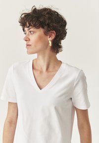 Camiseta de algodón blanca con escote en V y mangas cortas, textura suave. Pendientes abstractos dorados, que añaden contraste visual.