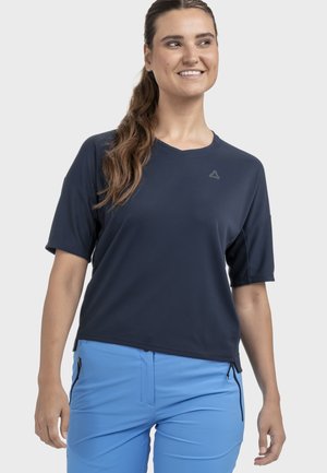 Frau lächelt, trägt ein marineblaues kurzärmeliges Hemd und hellblaue Hosen mit seitlichen Reißverschlusstaschen, steht vor einem einfarbigen Hintergrund.