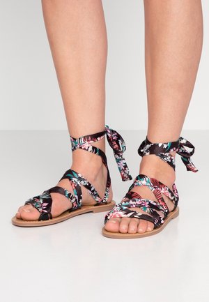 Sandaletter med blomsterprint, flade med sorte stofremme, åben tå og bindebånd ved anklen. Lys tan ydersål, designet til afslappet brug.