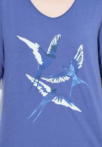 Tres golondrinas estilizadas en azul y blanco con las alas extendidas impresas en la parte delantera de una camiseta de manga corta morada.
